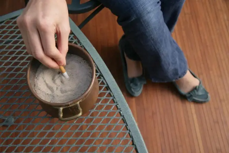 A person sitting on a chair outside, snuffing out a cigarette in an ashtray on a metal table.