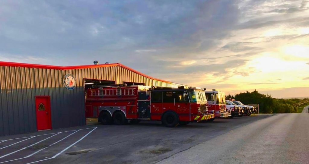 Four fire trucks are parked outside a fire station at sunrise or sunset, with the sky showing clouds and warm light. The fire station has a gray and red exterior with an emblem above the door.