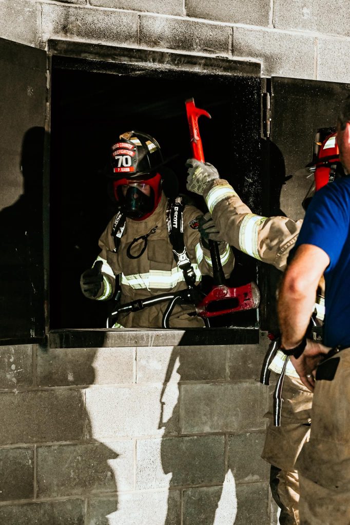 A firefighter in full gear and helmet exits a dark window holding a red axe, while another firefighter and a person in a blue shirt stand nearby against a cinder block wall.