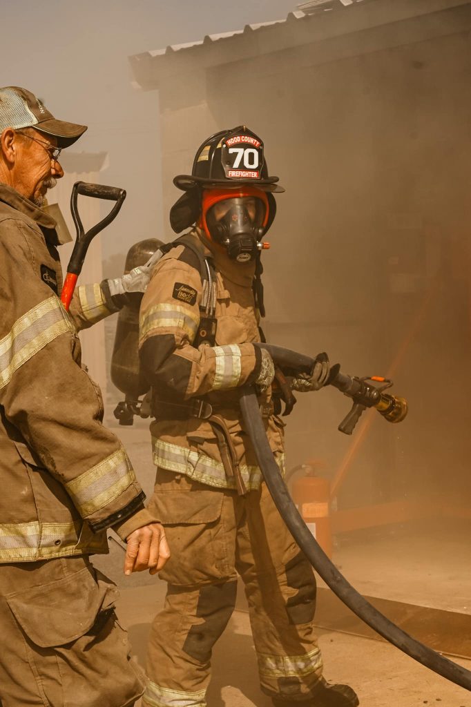 Two firefighters in protective gear and helmets stand amid smoke; one holds a shovel while the other, wearing a breathing mask, carries a fire hose, preparing to tackle a fire near a building.