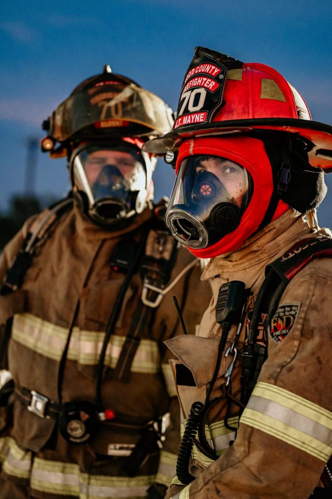 Two firefighters in full gear, including helmets, face masks, and protective suits, stand side by side outdoors at dusk, prepared for emergency response.