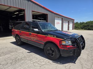 A black and red fire department SUV labeled “Squad 70” is parked outside a metal garage building with several white garage doors on a sunny day.