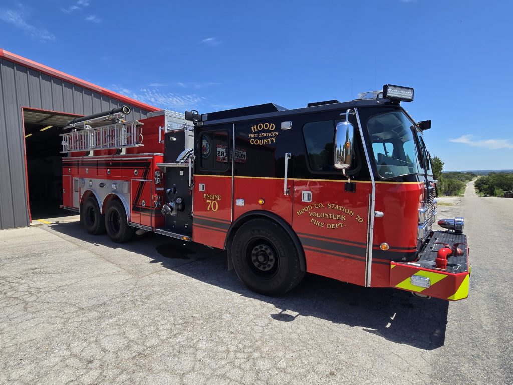 A large red fire truck labeled "Hood Co. Station 70 Volunteer Fire Dept." is parked outside a metal building on a sunny day, with equipment and ladders visible on top of the truck.