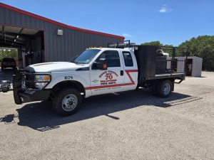 A white flatbed fire truck labeled "Hood County Volunteer Fire Dept" is parked outside a metal fire station building on a sunny day. The truck has emergency lights and equipment on its bed.