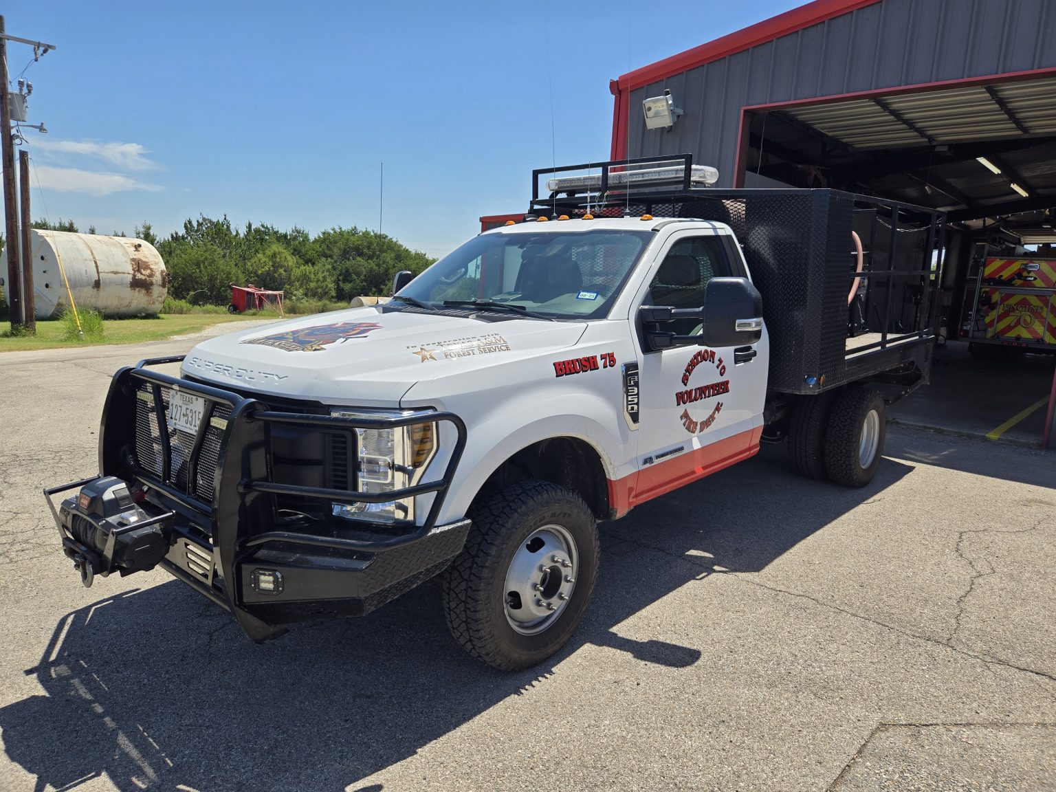 A white fire department brush truck labeled "BRUSH 75" is parked outside a fire station, equipped with off-road tires, a front grille guard, and a black utility bed for firefighting equipment.