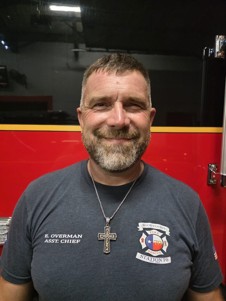 A man with short hair and a beard, wearing a navy blue firefighter shirt labeled "E. Overman Asst. Chief" and a cross necklace, smiles in front of a red fire truck.