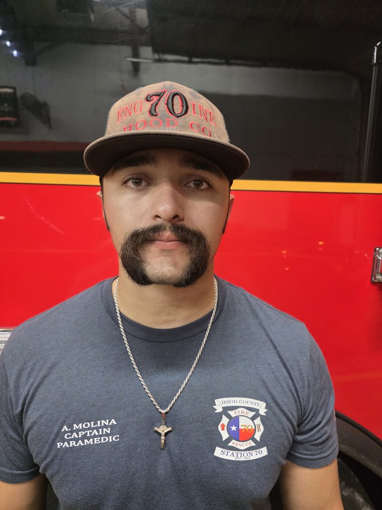 A man with a mustache and chin beard wears a "Hood Co 70 Line" cap, a blue t-shirt labeled "A. Molina Captain Paramedic," and a necklace with a cross, standing in front of a red vehicle.