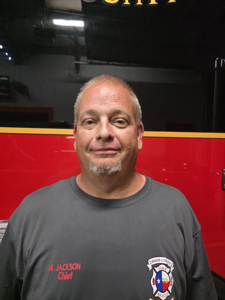 A middle-aged man with short gray hair and a goatee stands in front of a red fire truck, wearing a gray shirt with a fire department logo and the words "M. Jackson Chief" embroidered on it.
