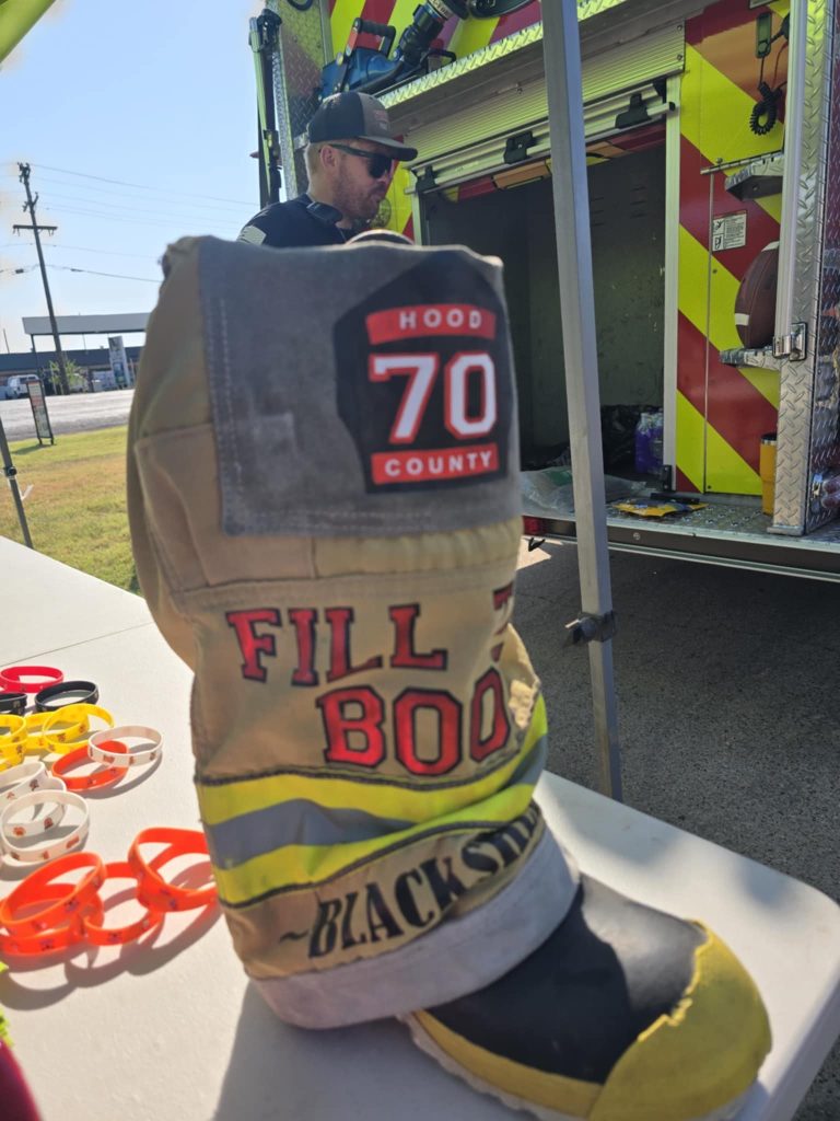 A large firefighter boot labeled “Fill the Boot” and “Hood County 70” sits on a table beside colorful wristbands, with a fire truck and a person in the background.