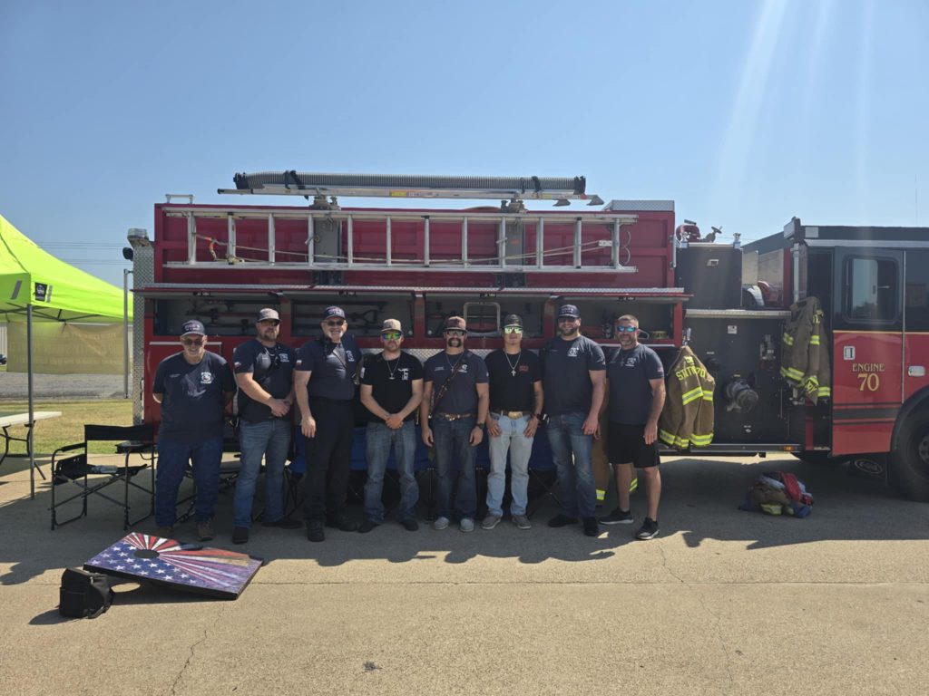 Nine firefighters in navy shirts and jeans or shorts stand in front of a red fire truck on a sunny day. A cornhole board and fire gear are on the ground nearby. A green tent canopy is visible on the left.