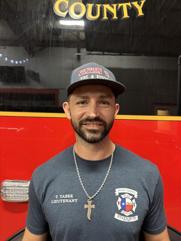 A man in a blue Station 70 Fire & Rescue shirt and hat stands in front of a red fire truck. He has a beard, wears a cross necklace, and is smiling at the camera.