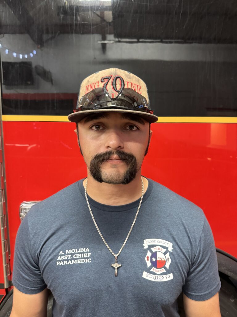 A man with a mustache, wearing a tan cap, sunglasses, and a blue T-shirt labeled “A. Molina Asst. Chief Paramedic,” stands in front of a red fire truck.