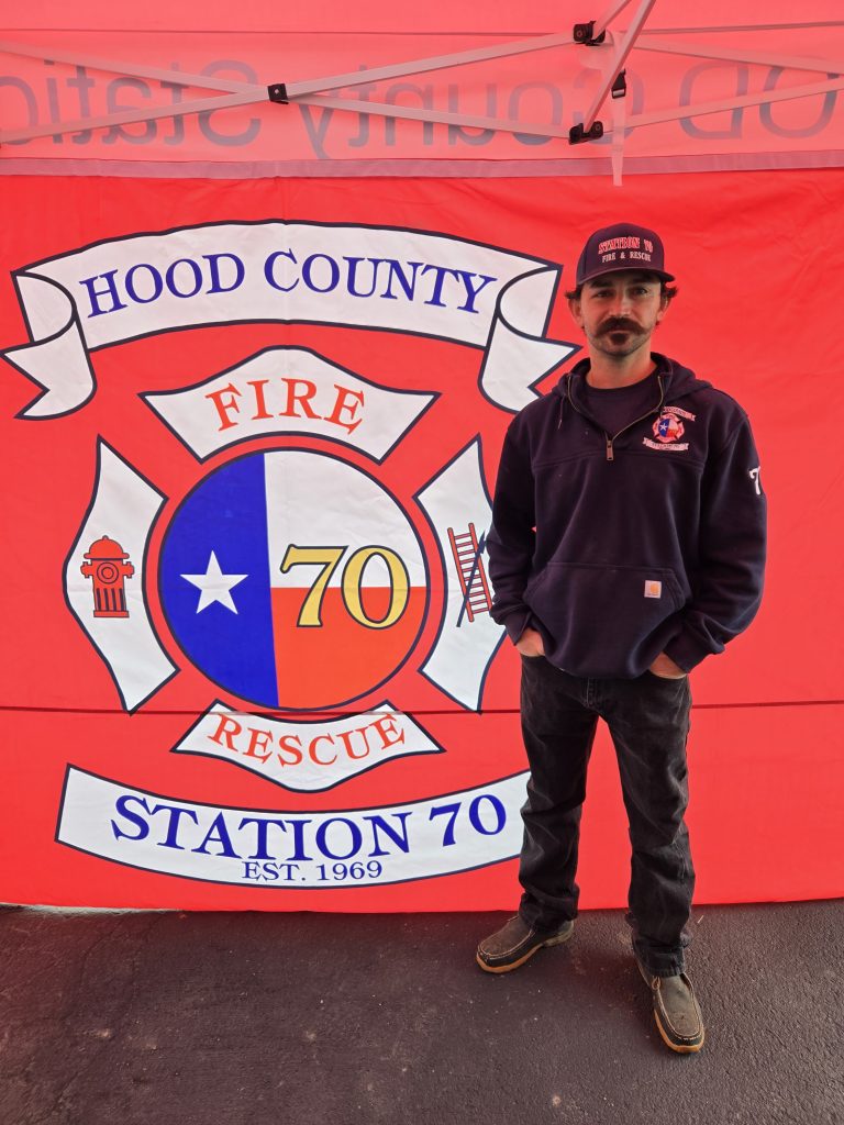 A man wearing a navy hoodie and cap stands in front of a red banner for Hood County Fire Rescue Station 70, featuring a Texas-themed emblem and fire department symbols.