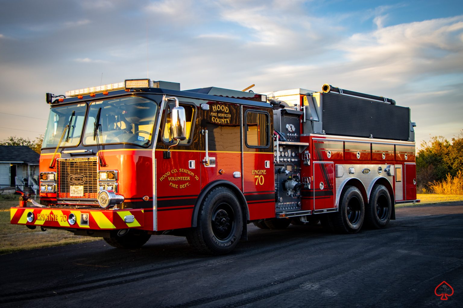 A bright red fire truck labeled "Engine 70" and "Hood County" is parked on a paved road with grass and trees in the background, illuminated by golden sunlight.