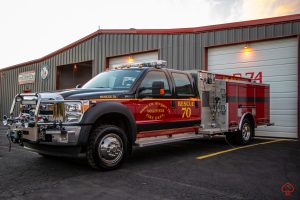 A red and black rescue fire truck labeled "Rescue 70" is parked in front of a metal fire station building with a large white garage door and the number 74 on it.