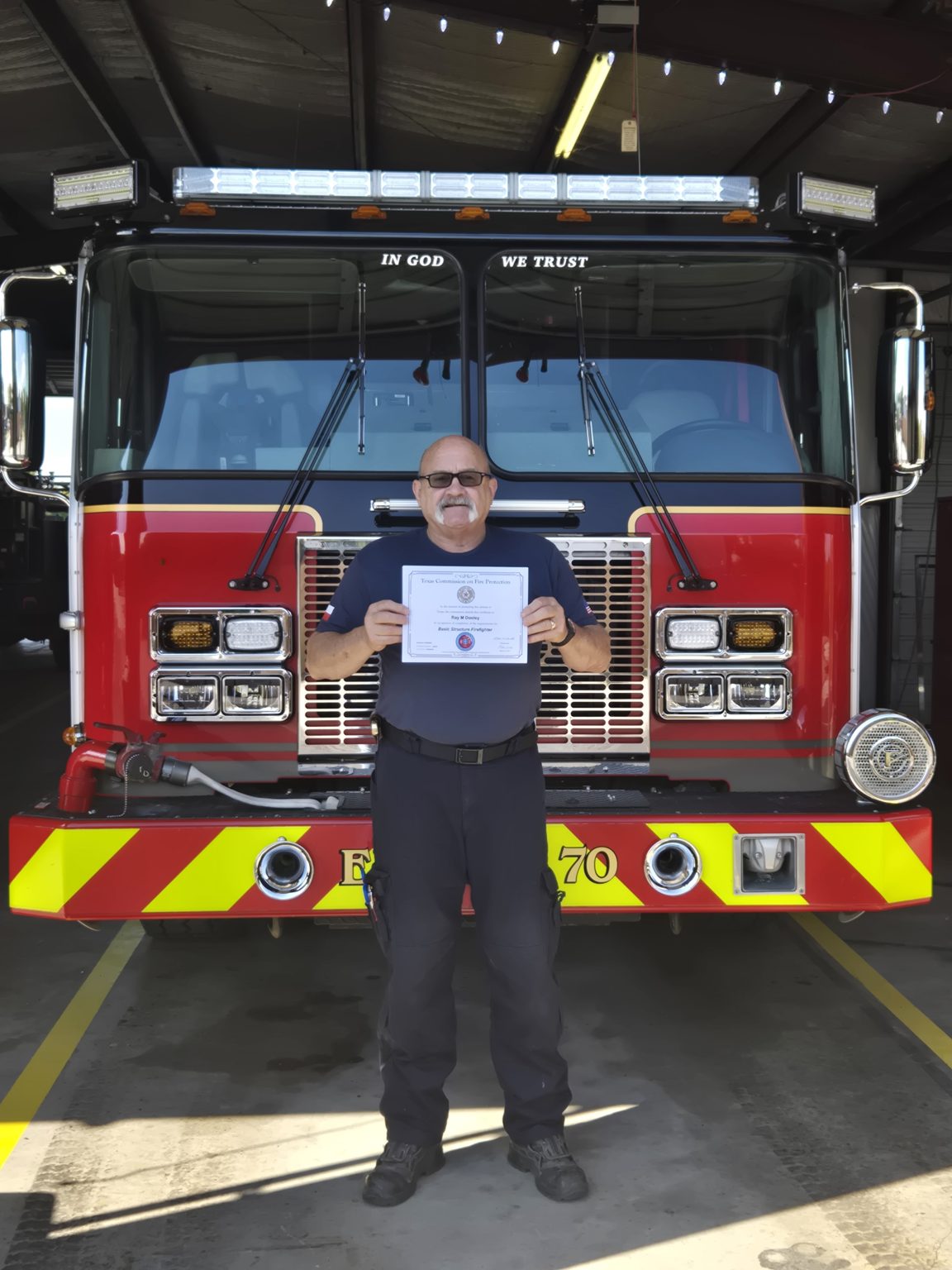 A man in a fire department uniform stands in front of a fire engine, holding a certificate and smiling for the camera inside a fire station.