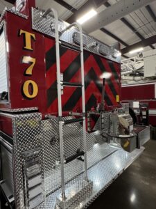 Close-up of the rear of a red fire truck labeled “T70,” with a metal ladder, diamond-plate platform, and black and red chevron stripes. The truck is inside a building under fluorescent lights.