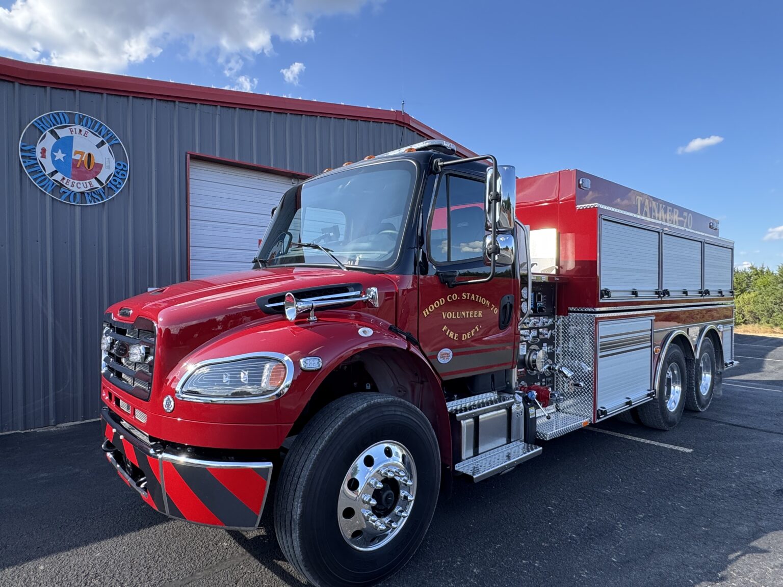 A shiny red fire truck is parked outside a metal building with a circular sign. The sky is blue with a few clouds.